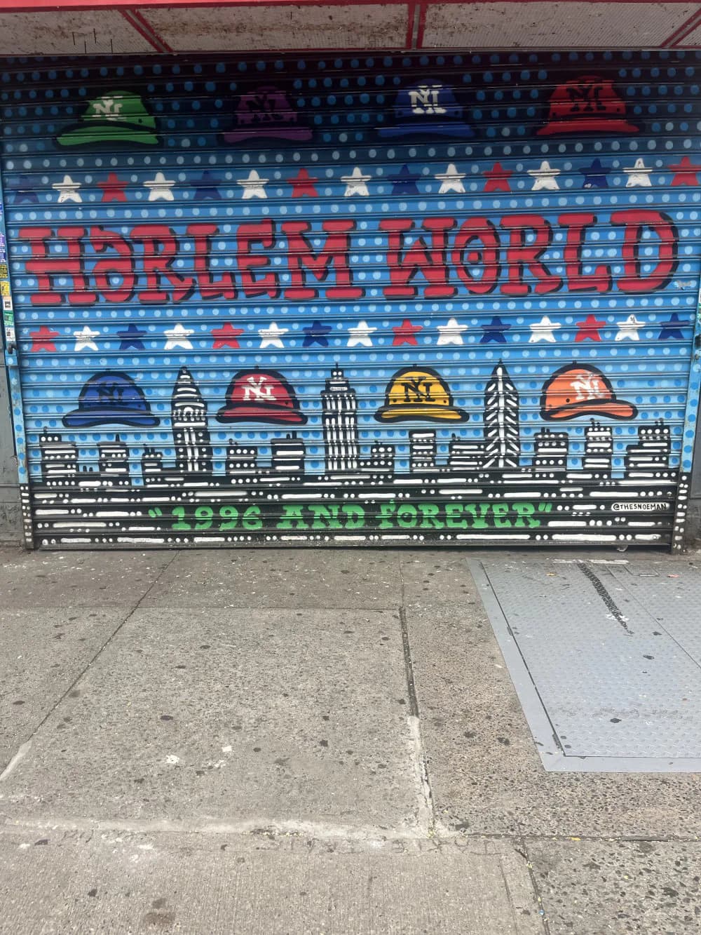 Mural on blue corrugated metal shutter featuring 'Harlem World' text in red letters with colorful hard hats and NYC skyline, stars and stripes pattern.