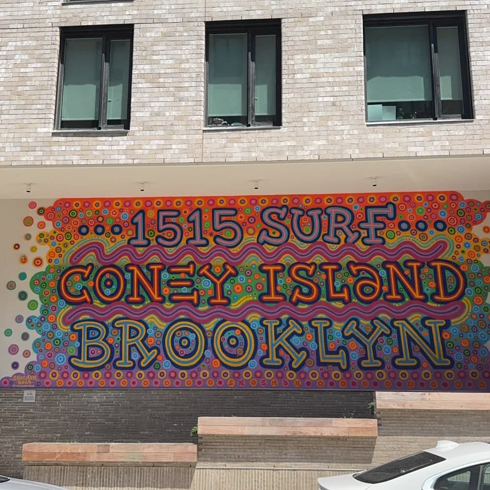 Mural on building facade reading '1515 Surf Coney Island Brooklyn' in outlined letters with rainbow dot pattern background in reds, blues, and greens.