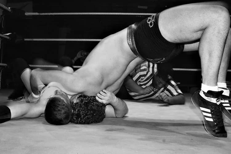 Black and white photograph of a shirtless male figure in an inverted pose on a wrestling ring, wearing black shorts and sneakers.