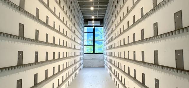 Photograph of a modern institutional corridor with tall white walls lined with uniform recessed storage compartments and centered blue window opening.