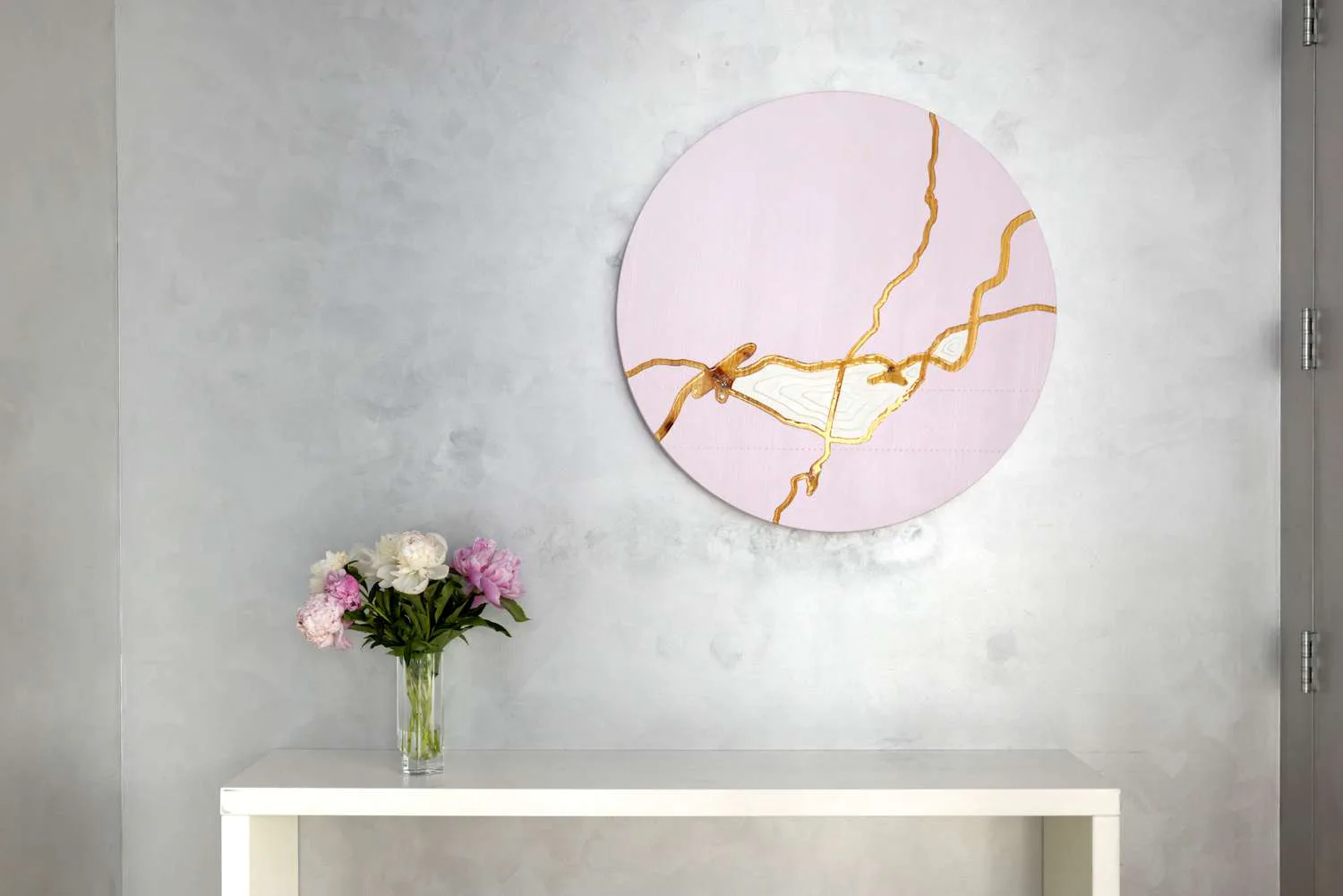 Gallery wall display: pale pink circular artwork with gold branch-like lines; white shelf below with pink flowers in clear vase.