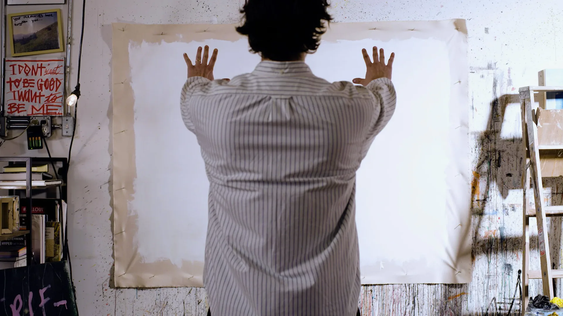 Figure with arms raised viewed from behind, painted in muted tones on weathered white wall surface with visible studio materials nearby.