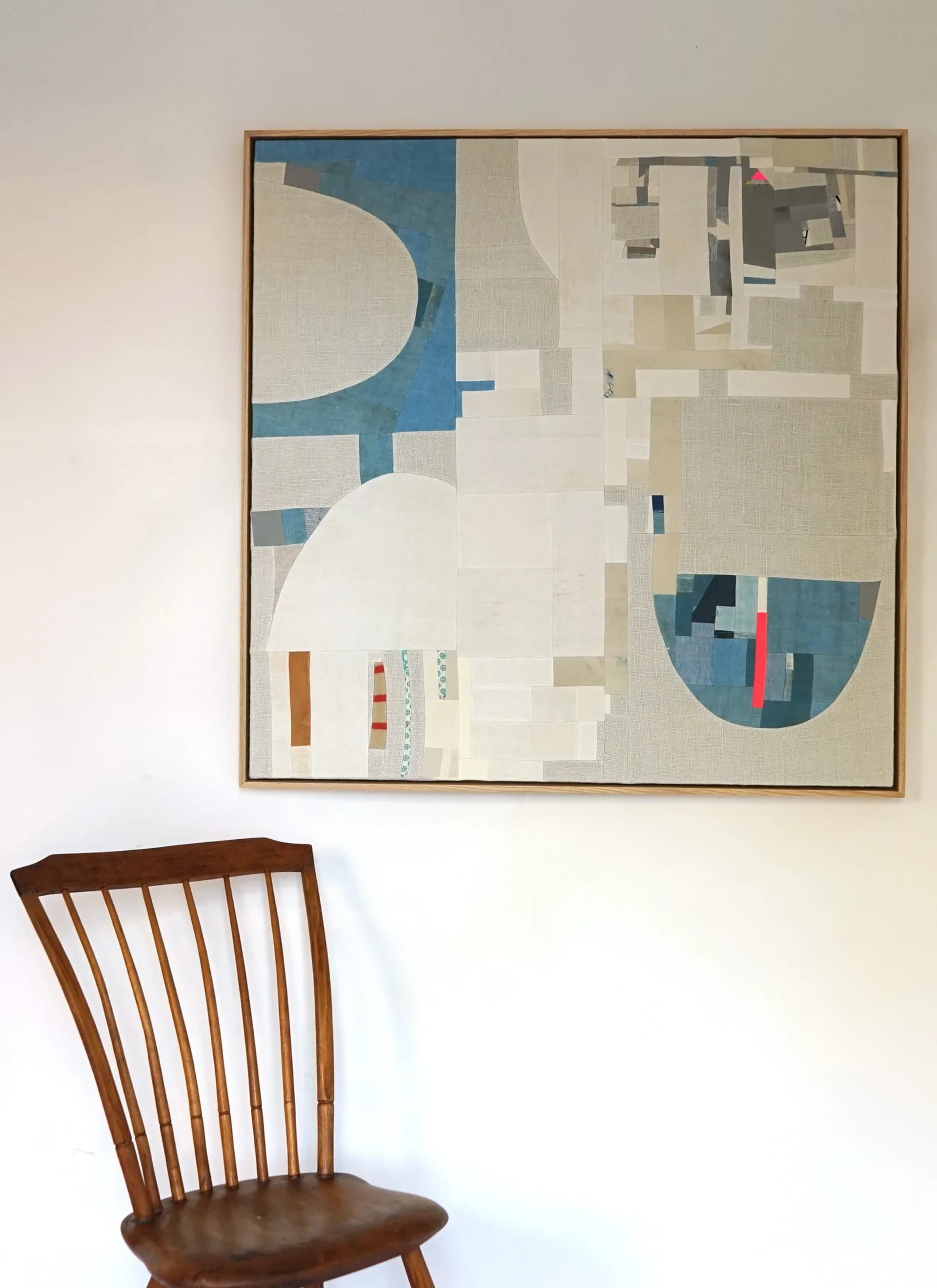 Framed abstract textile artwork displayed on white gallery wall above a wooden mid-century chair, featuring blue, cream, and geometric shapes.