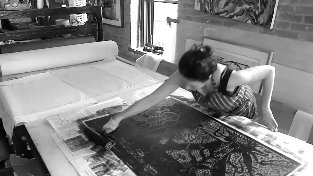 Black and white photograph of an artist working in a print studio, leaning over a large print on a work table with equipment visible in background.