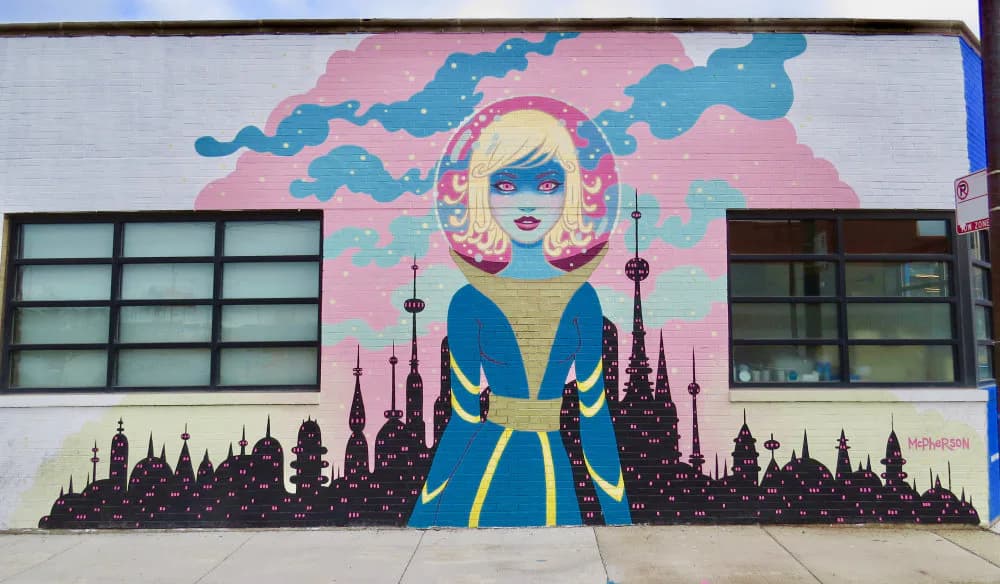 Mural of a stylized woman with blonde hair and blue skin wearing a blue and gold coat, standing before a silhouetted cityscape with pink and teal sky.