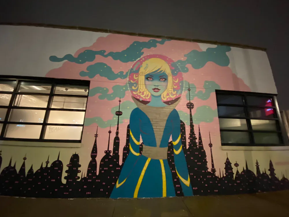 Mural of a stylized woman with blonde hair and blue skin in blue and gold coat before silhouetted city skyline, photographed at dusk with interior lighting visible.