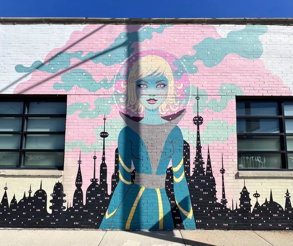 Mural featuring stylized woman with blonde hair, blue skin, blue and gold coat standing before black silhouetted cityscape with pink and teal sky, in bright daylight.