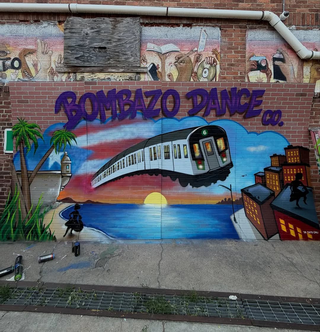 Brick wall mural titled 'SOMBAZO DANCE' featuring a stylized train flying through sunset sky with palm trees, water, and buildings below in blue, orange, and pink tones.