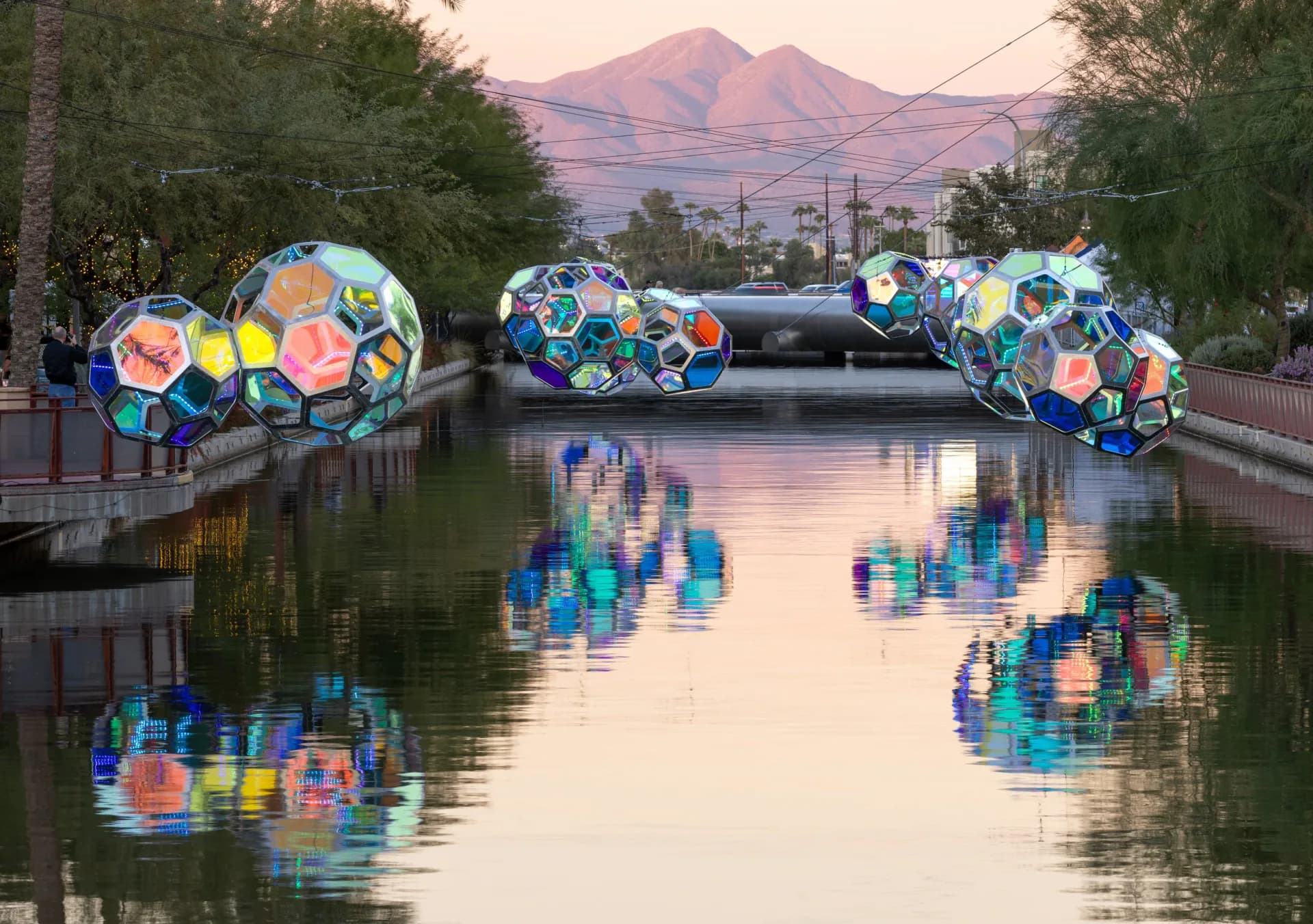 Iridescent geodesic sphere sculptures suspended over a canal at sunset with mountain backdrop