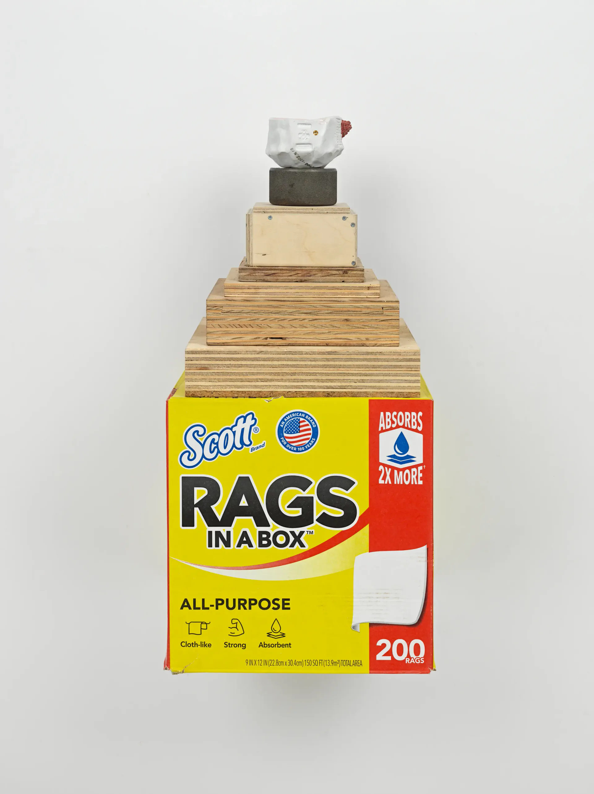 Stacked assemblage sculpture: yellow Scott Rags box base topped with wooden blocks, metal box, and small objects, creating a tower composition.
