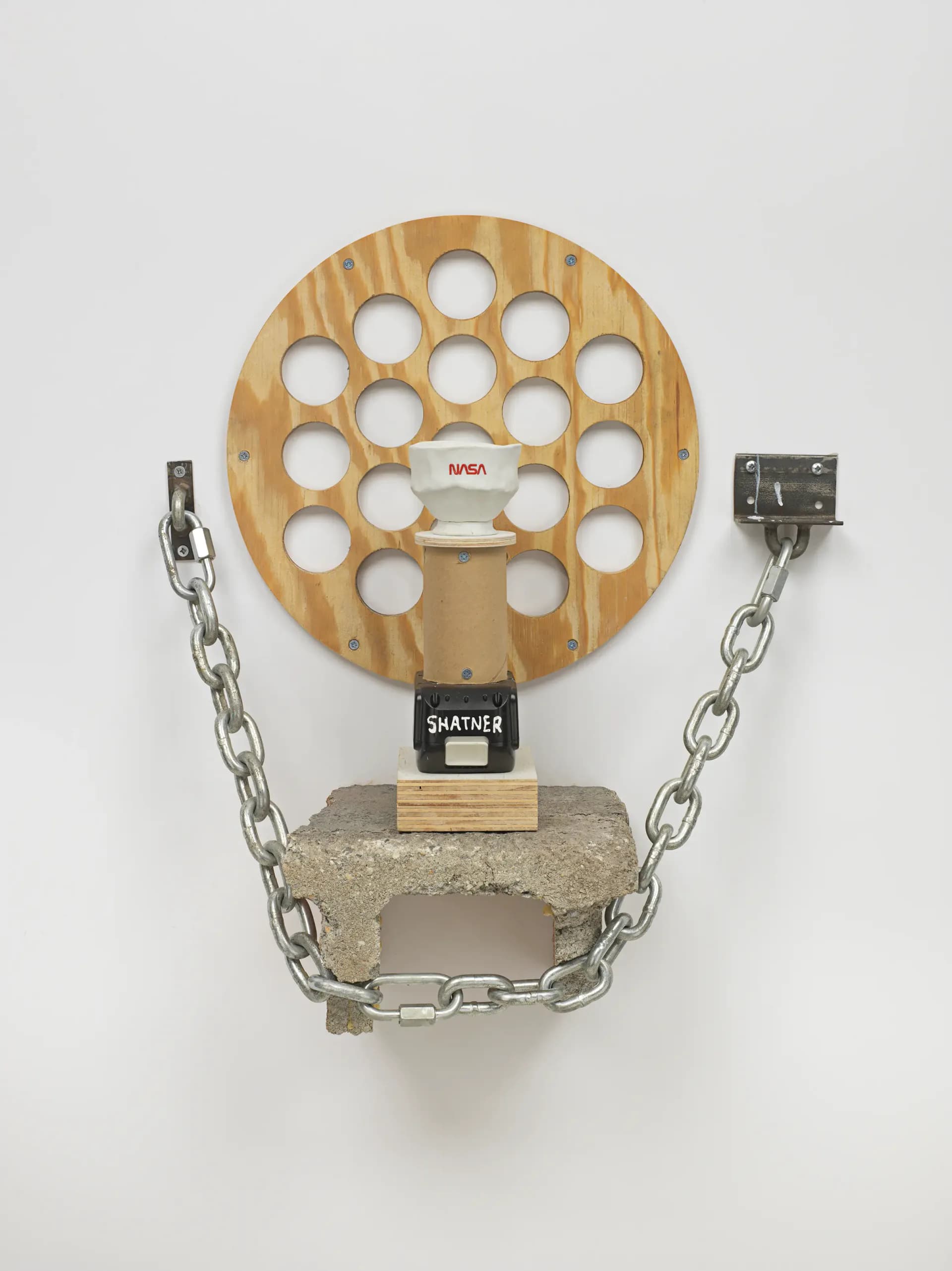 Wooden circular perforated disk with NASA branding, suspended by metal chains and brackets from a concrete base.