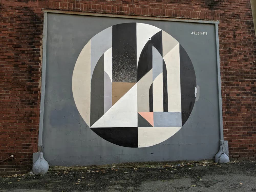 Mural within circular composition combining geometric shapes in black, white, beige, and muted blue-gray tones on brick wall.