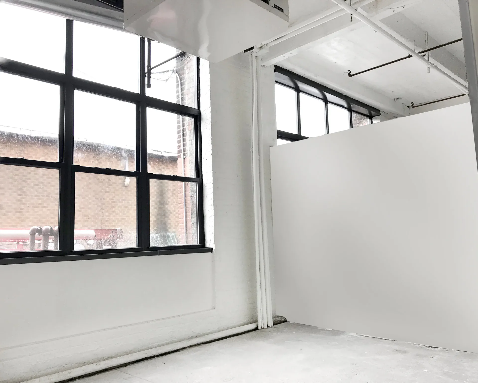 Interior of Trestle Art Space with white walls, industrial ceiling, large black-framed window overlooking a brick building, and polished concrete floor.