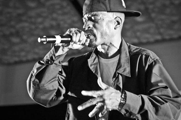 Black and white photograph of a performer singing into a microphone, wearing a cap and leather jacket with expressive hand gestures.