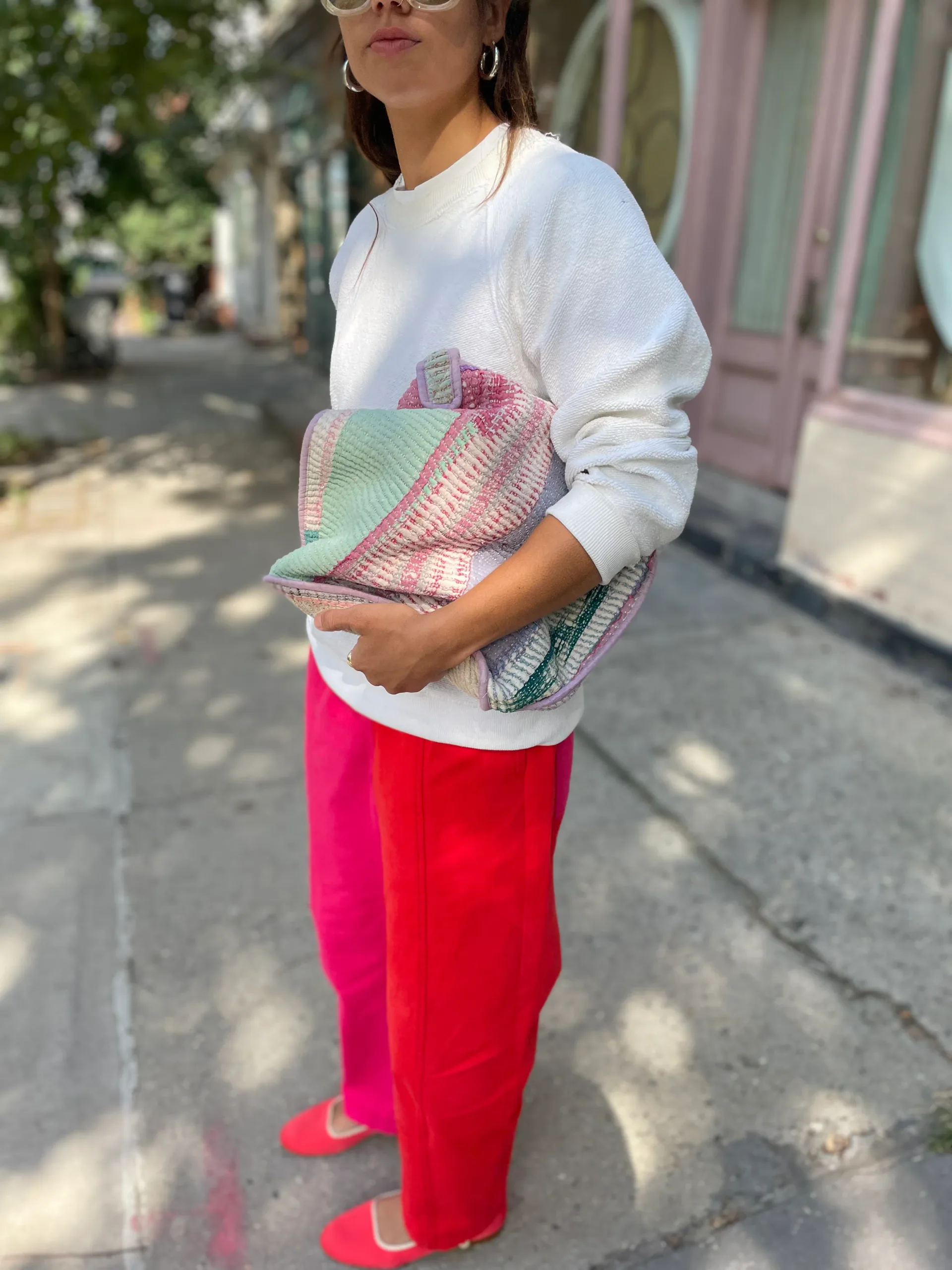 Person wearing a cream-colored long-sleeve Verity & Daughters sweatshirt, hot pink cotton pants, holding a multicolored woven bag, on a tree-lined street.