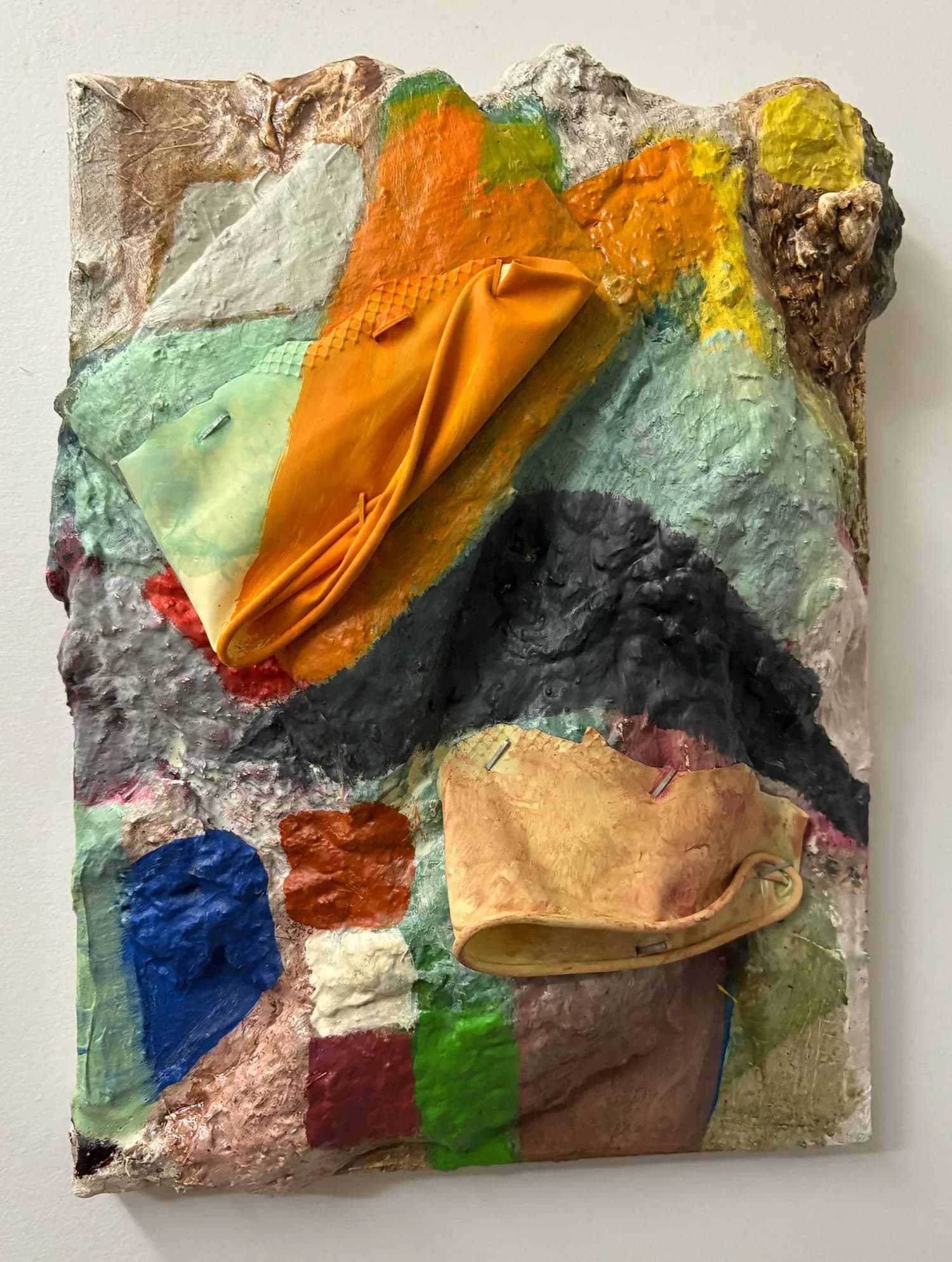Three-dimensional mixed-media artwork with sculptural fabric and paint elements in orange, green, yellow, black, and earth tones, featuring draped and dimensional forms against a pale textured surface.