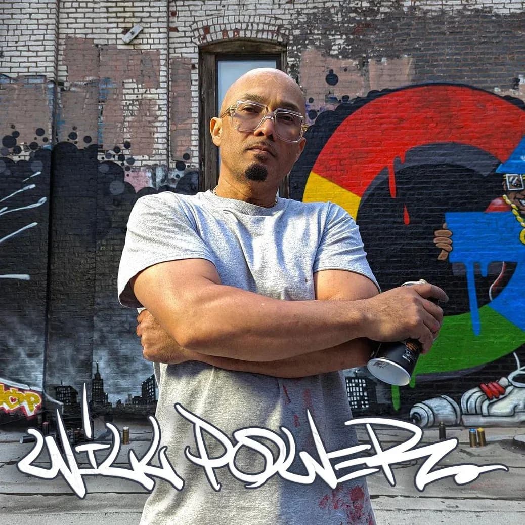 Man wearing glasses and light gray t-shirt posing with crossed arms in front of colorful graffiti mural background.
