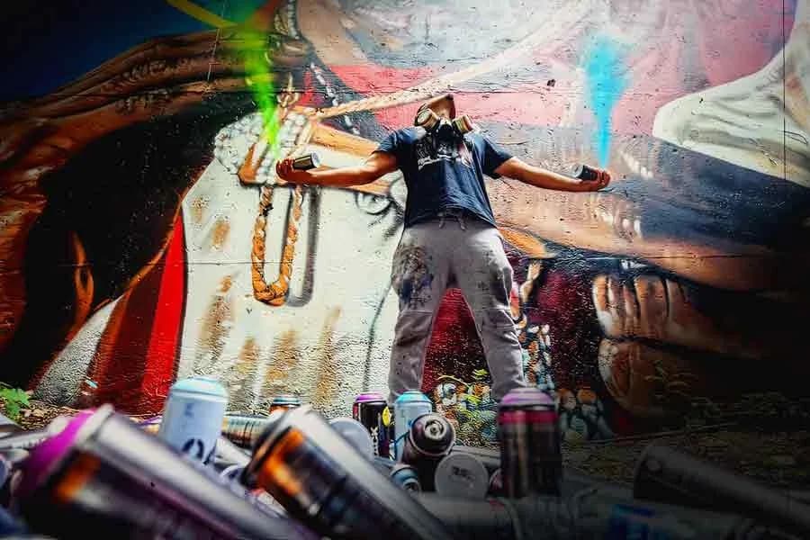 Mixed-media installation with person in dark clothing posed against large-scale painted figures and draped fabric, surrounded by spray paint cans.