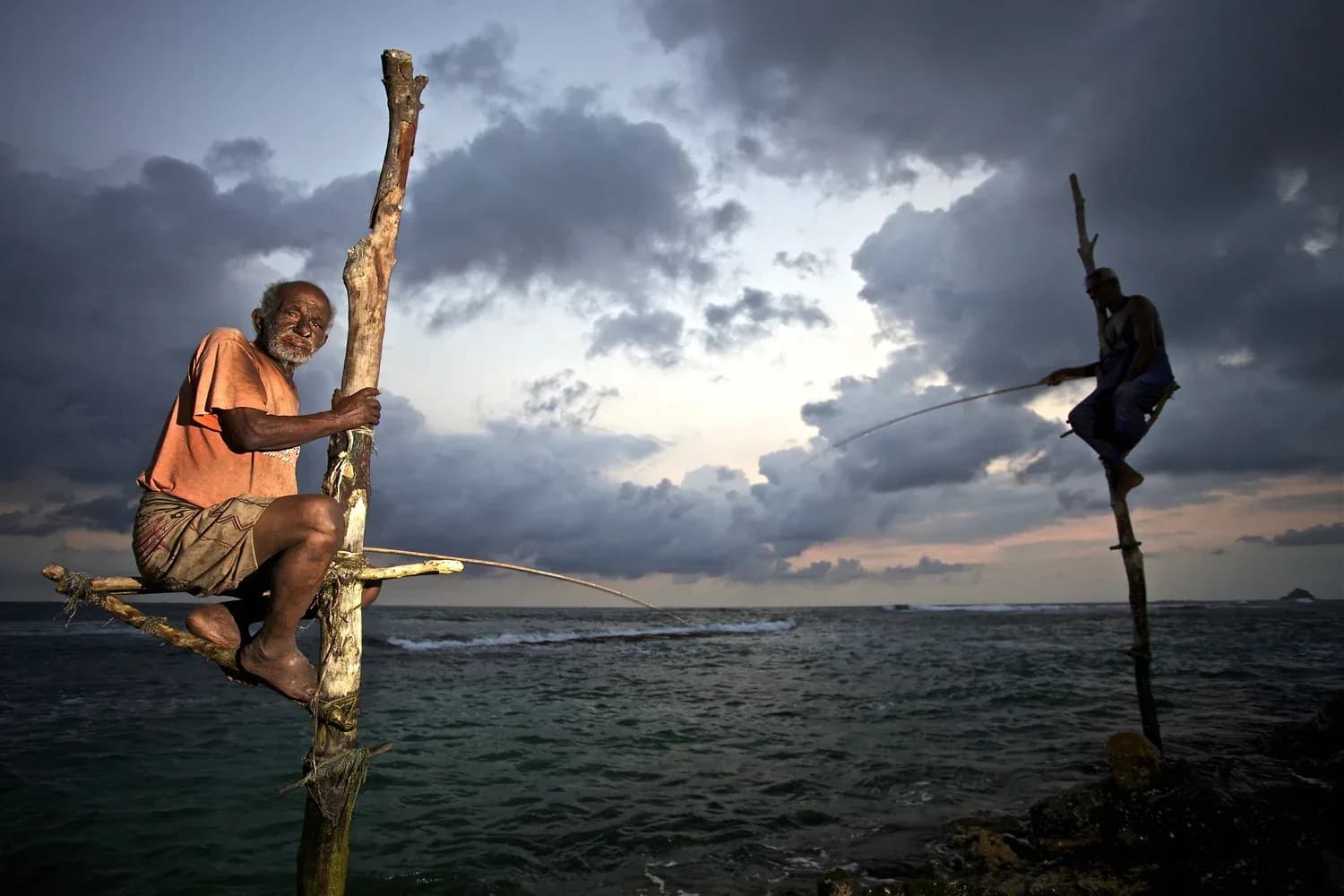 Sri Lankan stilt fishermen perched on poles over the ocean at dusk with dramatic storm clouds