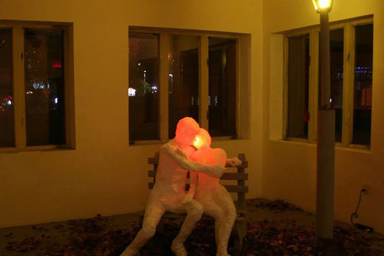 Mixed-media installation of a tan sculptural figure with glowing orange-red head, seated in yellow interior space with dark windows and carpet flooring.