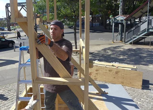 Photograph of man working on wooden frame construction outdoors in urban plaza with trees and buildings visible in background.