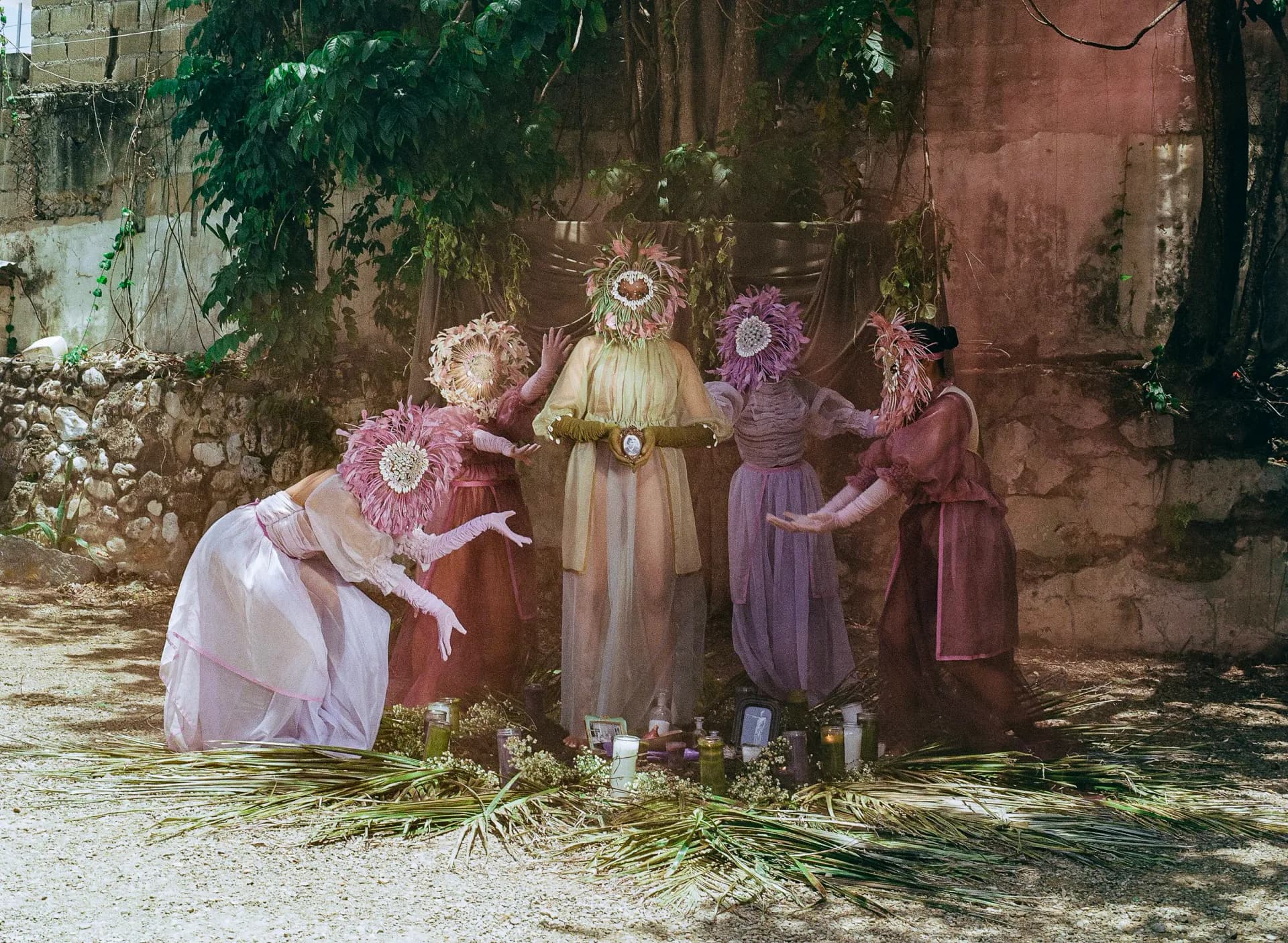 Four figures in flowing pastel dresses and large ornate circular headdresses pose in an aged stone courtyard installation by Yelaine Rodriguez.