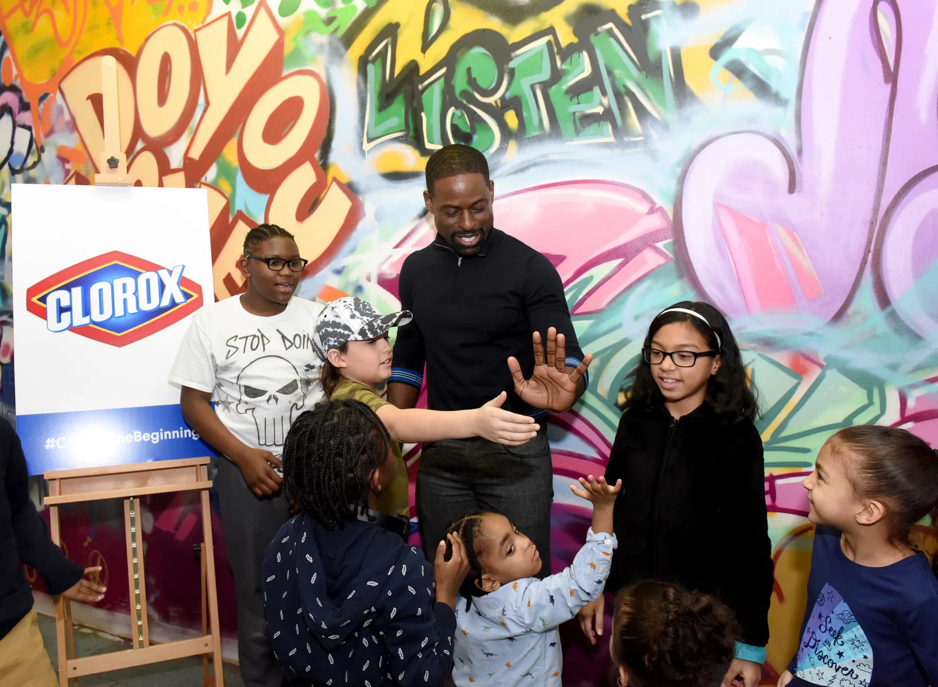 Vibrant mural with graffiti-style lettering in orange, green, and pink behind adults and children engaged in an educational activity with a Clorox branded display.