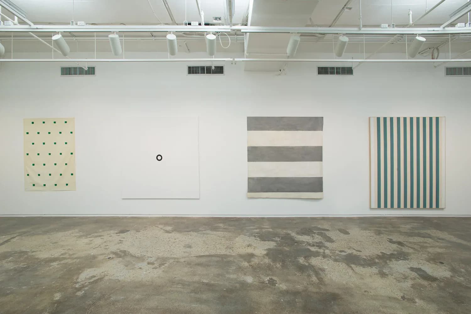 Gallery interior at 205 Hudson Gallery showing white walls with four abstract artworks: a yellow dotted piece, blank canvas, gray striped panels, and green striped composition on polished concrete floor.
