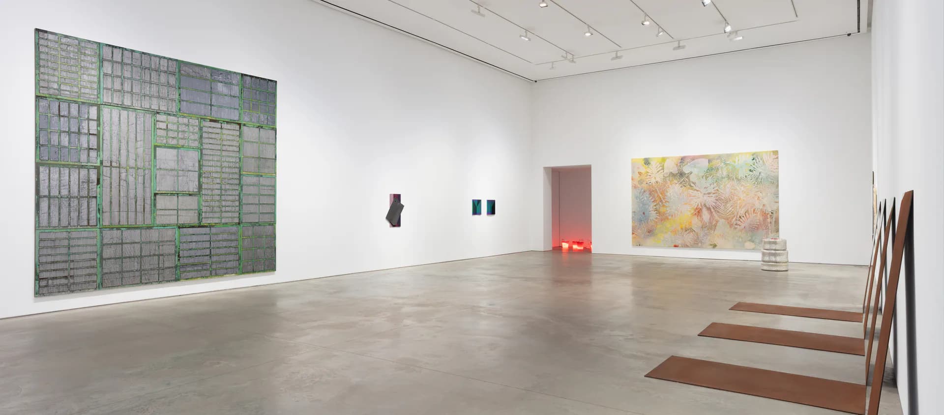 White gallery interior with polished concrete floor, featuring a large gridded abstract painting on left wall, small sculptures and paintings throughout, and striped floor element on right.