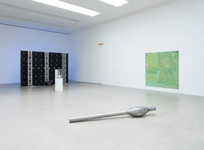 Gallery interior with white walls and fluorescent lighting, featuring a metallic sculptural element on floor, black patterned installation, and green abstract painting on wall.
