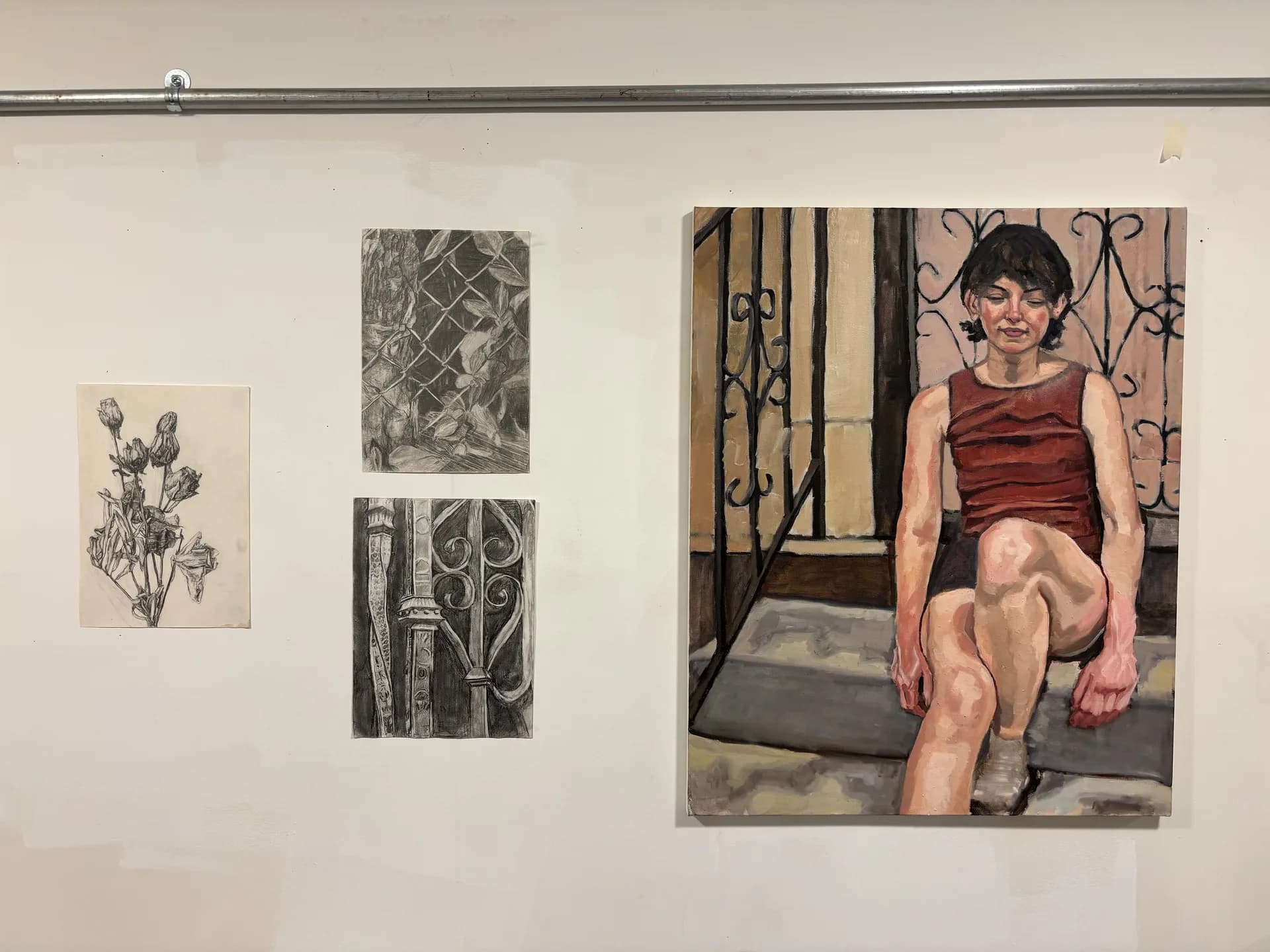 Gallery wall at 303 Woodbine Studio displaying two small charcoal drawings and a large painting of a seated woman in a red top near iron railings.