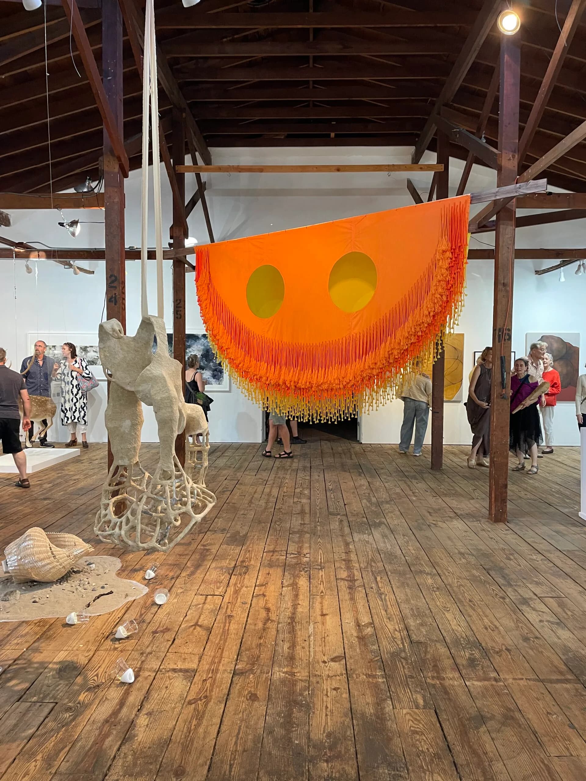 Interior of 4heads gallery showing wooden floor and exposed beam structure with visitors viewing artworks, including a large orange sculptural installation with fringed edges and eye-like circular openings.