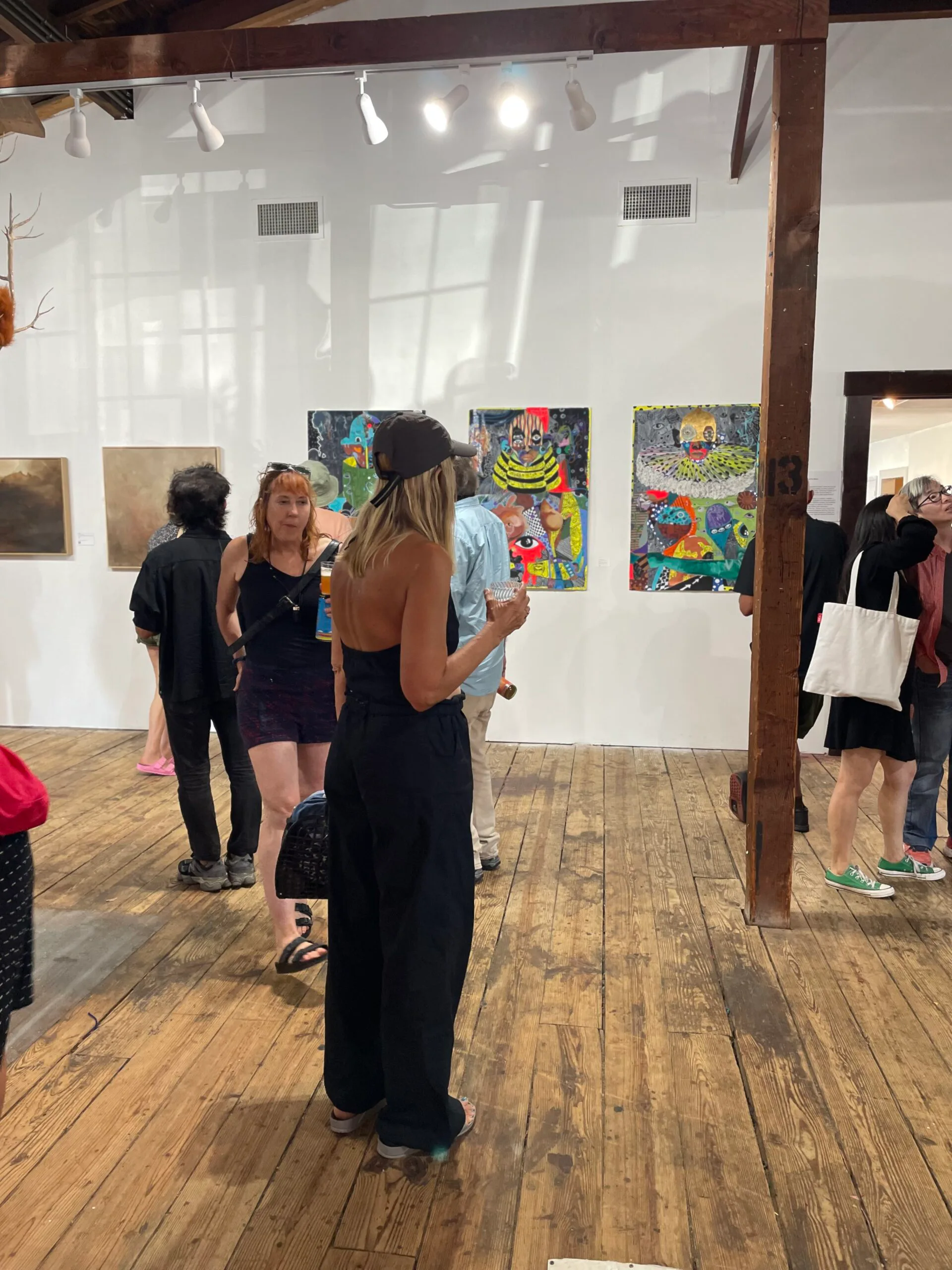 4heads gallery interior with wooden floors and structural columns; visitors viewing colorful paintings mounted on white walls, with natural light streaming through skylights.
