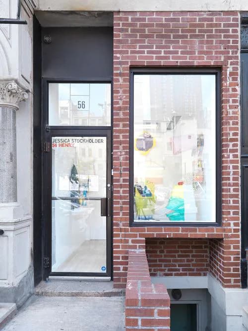 Storefront of 56 Henry gallery with black-framed glass door and window in red brick facade, interior visible with white walls and artwork.
