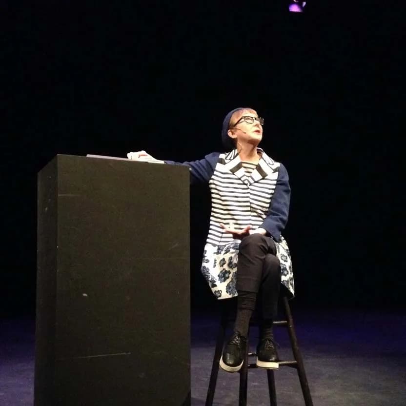Performance art or theatrical documentation: figure in striped shirt and patterned skirt seated on stool next to black rectangular structure on dark stage.