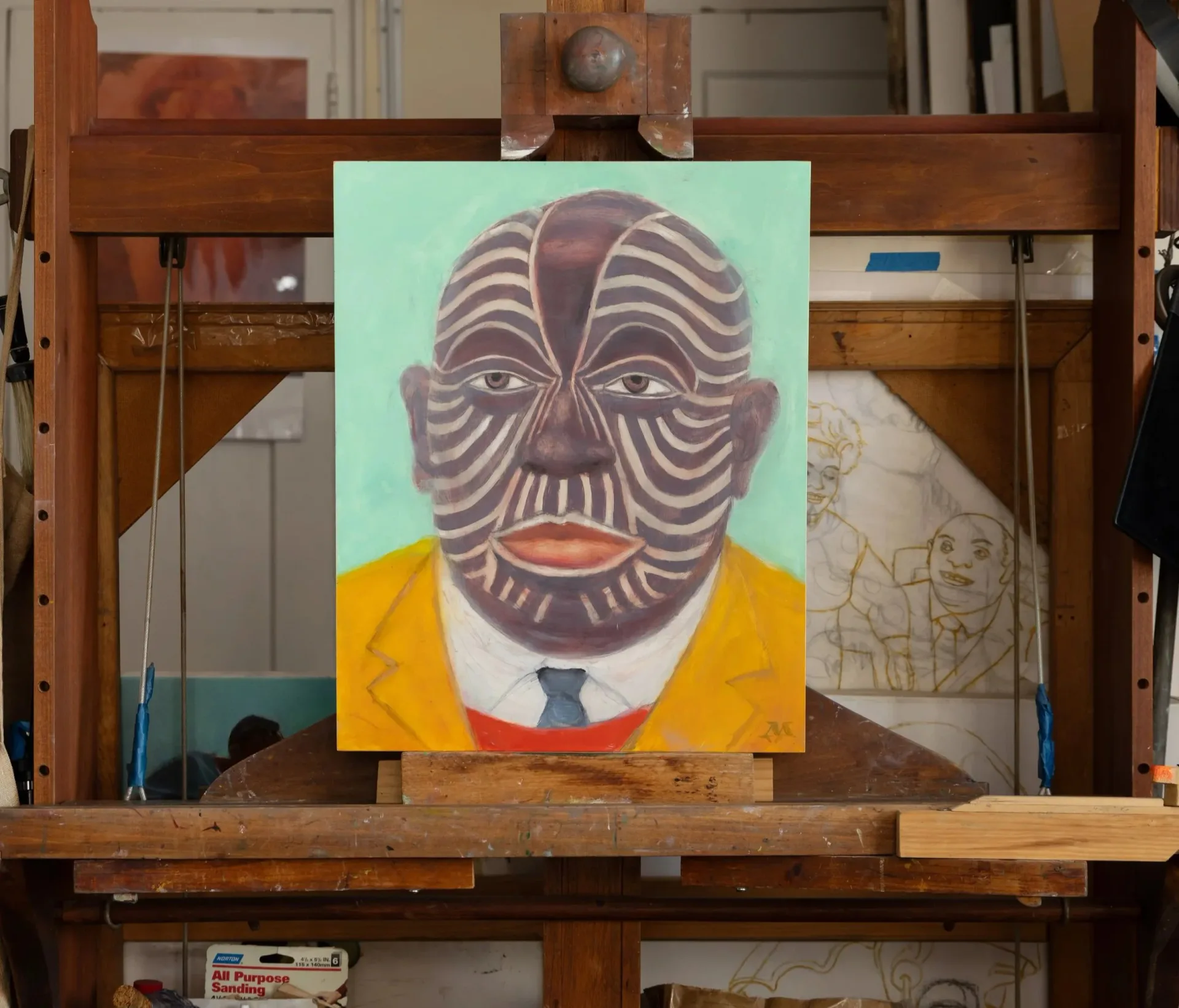 Wooden artist's easel in studio displaying portrait painting with geometric patterned face, yellow jacket, and tie against mint green background.