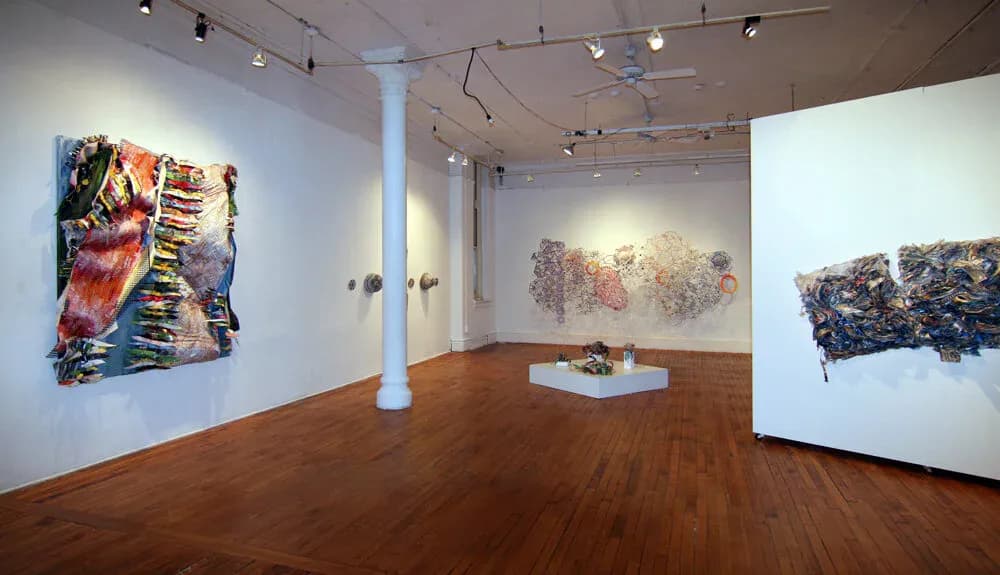 Modern gallery interior with wooden floors, white walls, and columns displaying abstract mixed-media artworks with bold colors and textural elements.