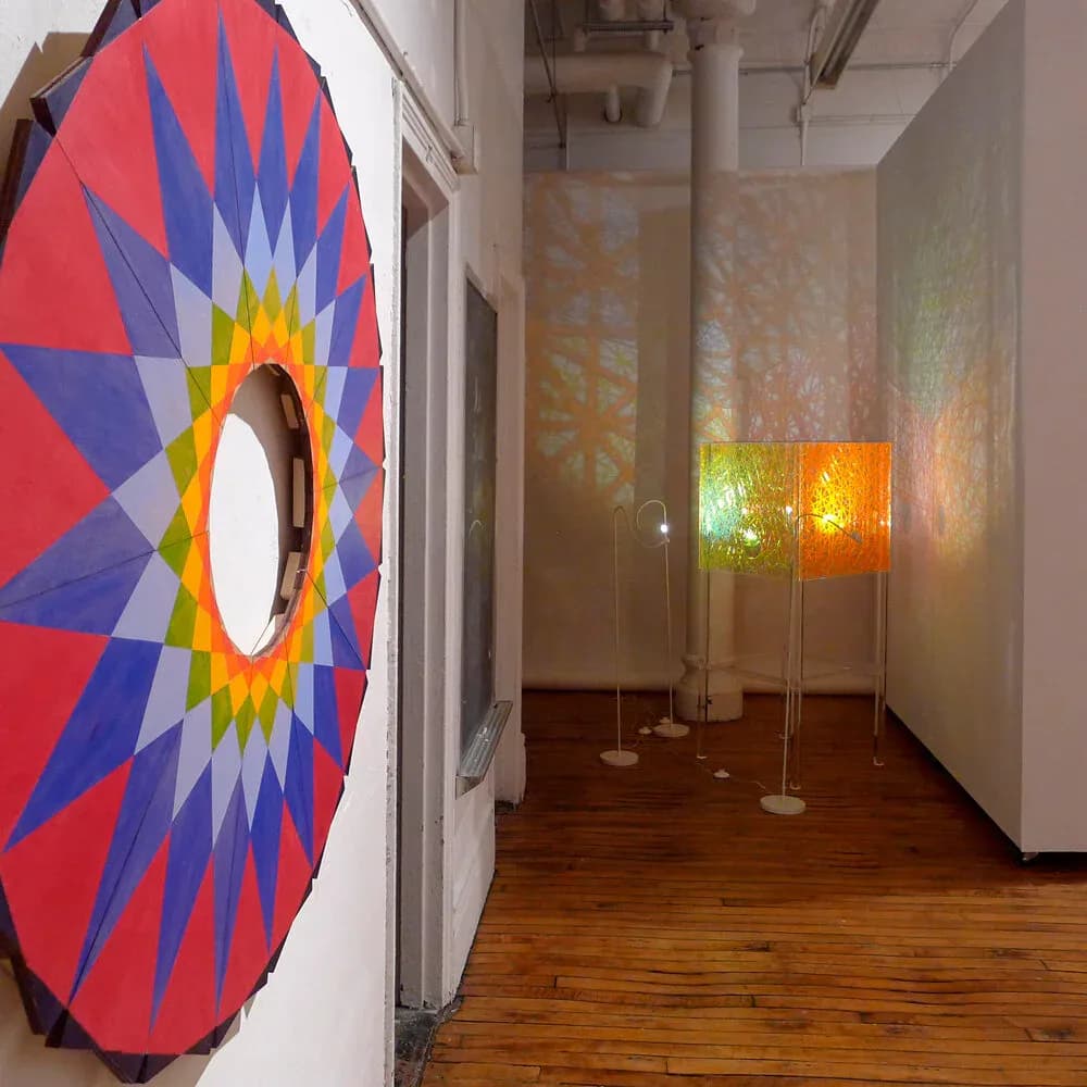 Gallery corridor with wooden flooring featuring a vibrant red and blue geometric circular artwork on the left and backlit amber-colored glass panels ahead.