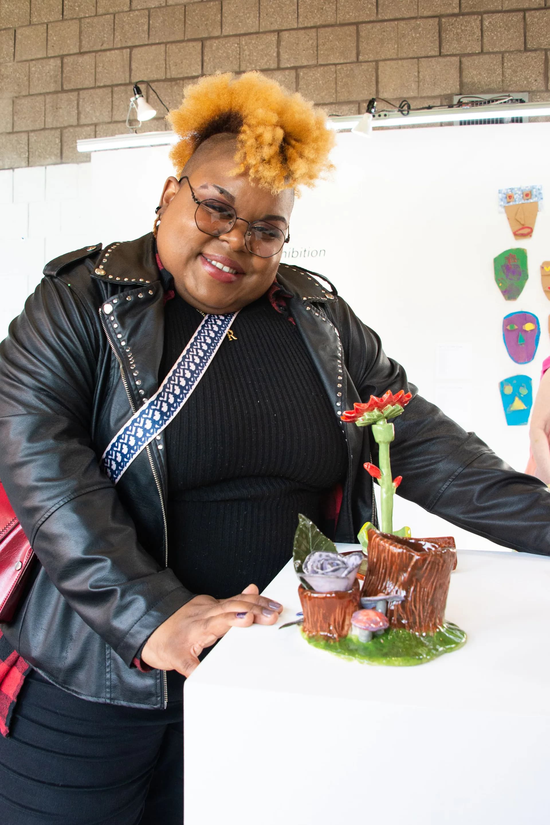 Person with bright yellow afro wearing black studded jacket and glasses, smiling at a table with handmade craft objects.
