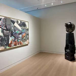 Modern gallery interior with light wood floors, white walls, framed painting on left wall, and black sculptural form on right.