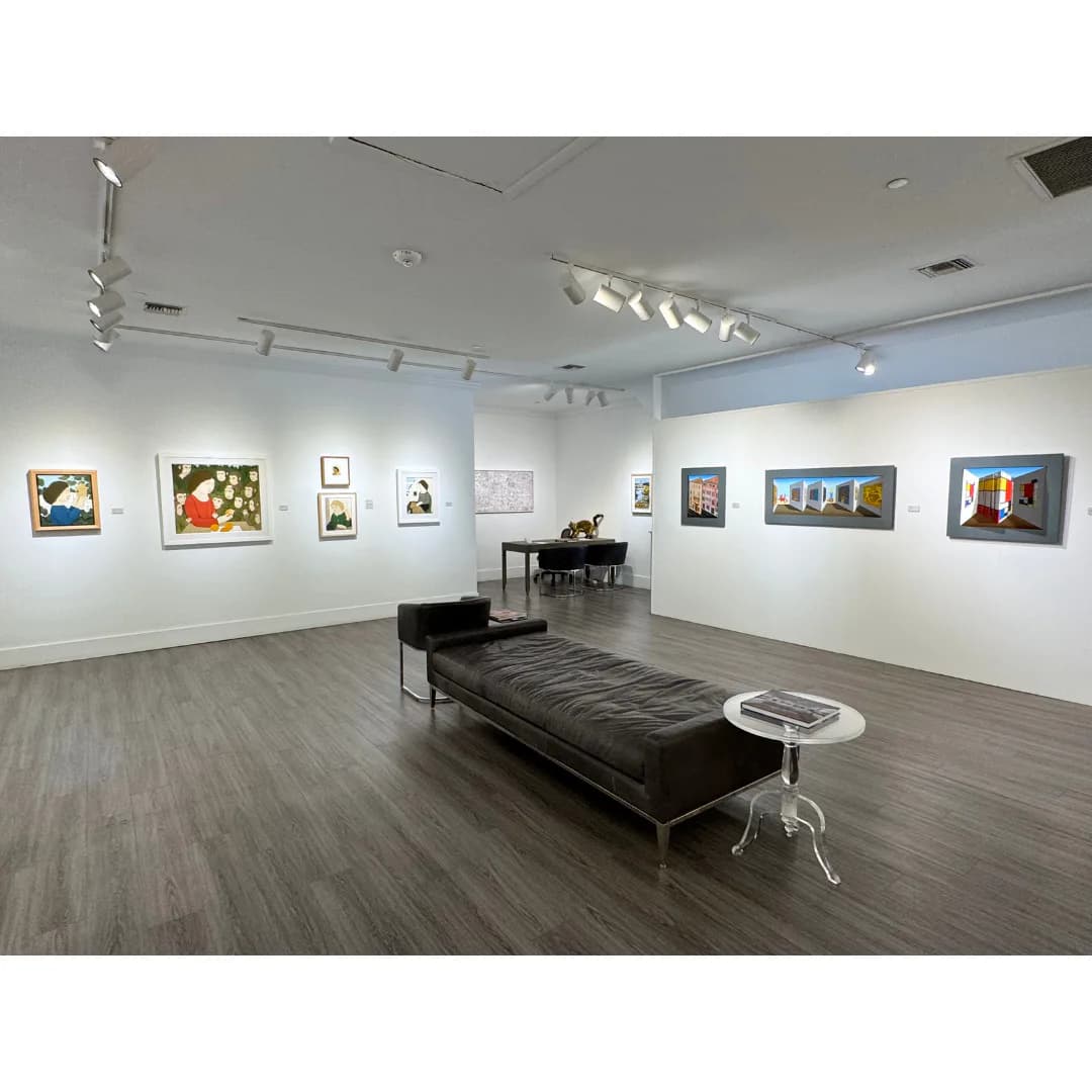Bright gallery interior with white walls, track lighting, dark piano, black bench, and colorful framed artworks arranged around the space.