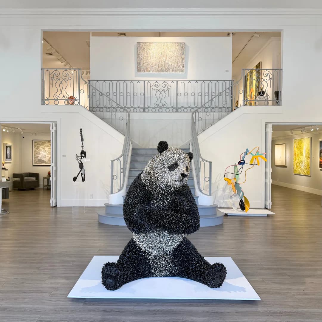 Multi-level gallery with dual staircases, white walls, and a large black textured sculptural figure seated in the center foreground.