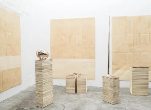 Gallery interior with white walls and pale wood panel artworks mounted on walls; sculptural stacks of wood blocks in natural finish arranged on polished concrete floor.