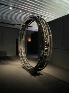 Gallery interior with a large circular black sculptural installation centered on a light floor, lit by overhead ceiling fixtures.