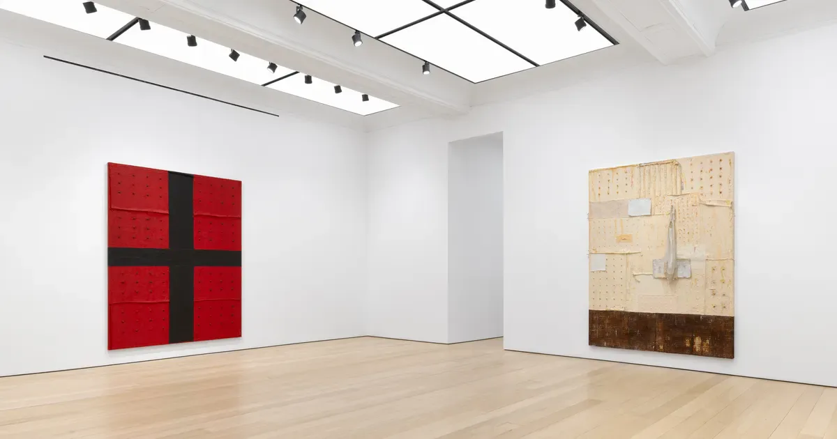 Minimalist gallery interior at Alexander Gray Associates with light wood flooring, white walls, and track lighting; a red geometric cross painting hangs left, a tan/beige mixed-media work hangs right.