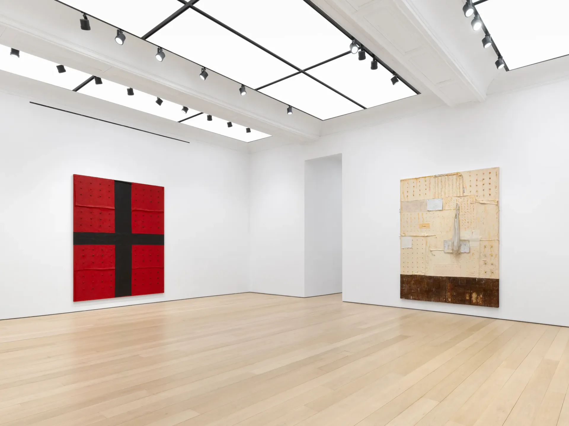 Gallery exhibition view at Alexander Gray Associates showing identical layout to previous image: red cross artwork on left wall, tan and brown mixed-media piece on right, light wood floor and contemporary lighting.