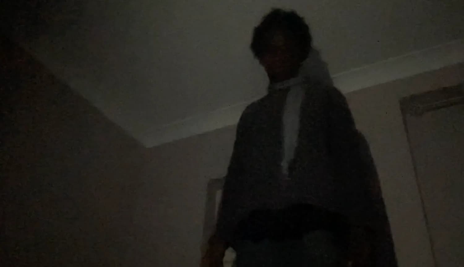 Dimly lit figure in dark clothing photographed indoors against neutral background; low image quality and unclear context.
