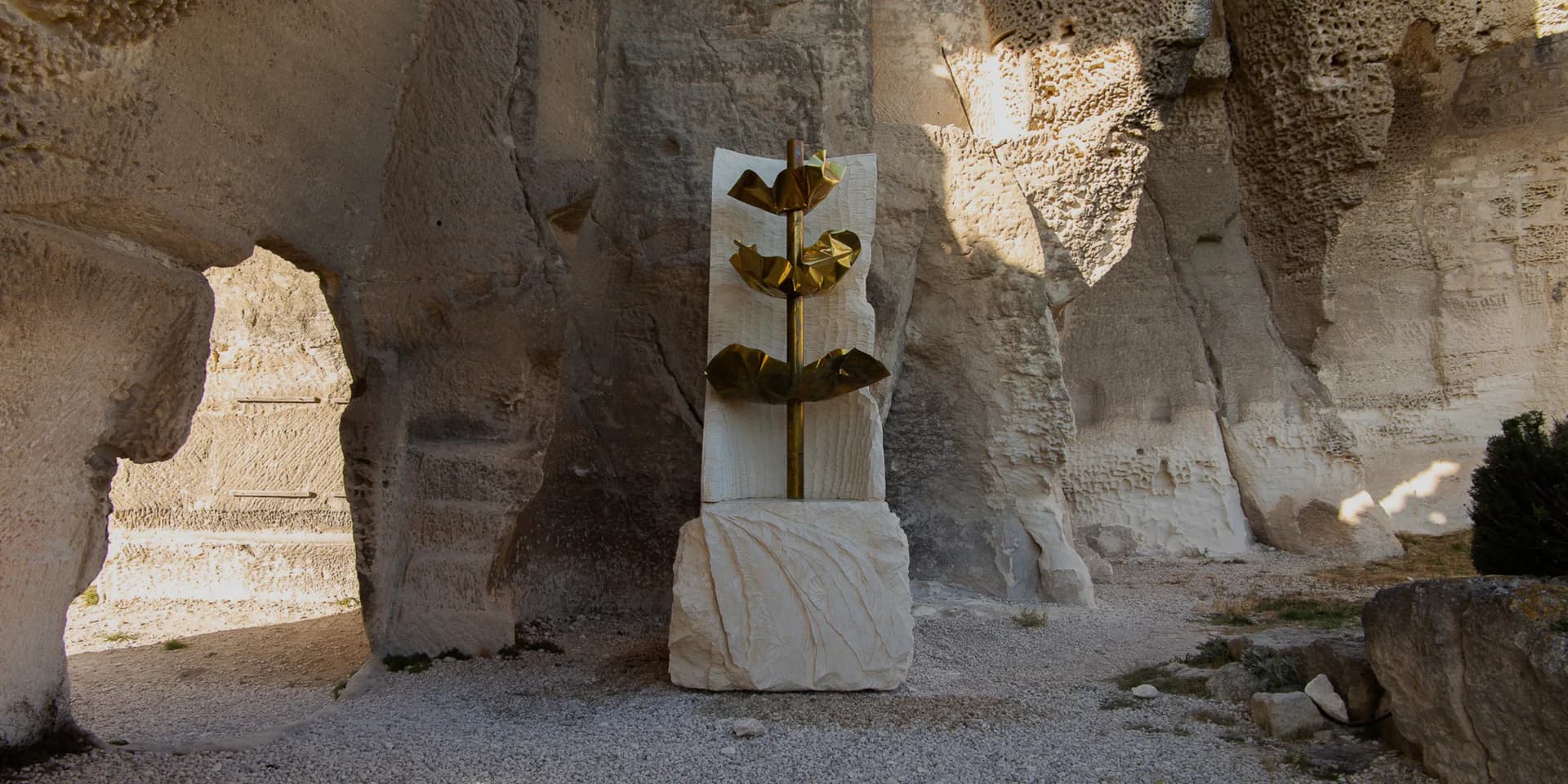 Installation within ancient stone ruins: white marble blocks arranged with central brass cross symbol, earthy minimalist composition.