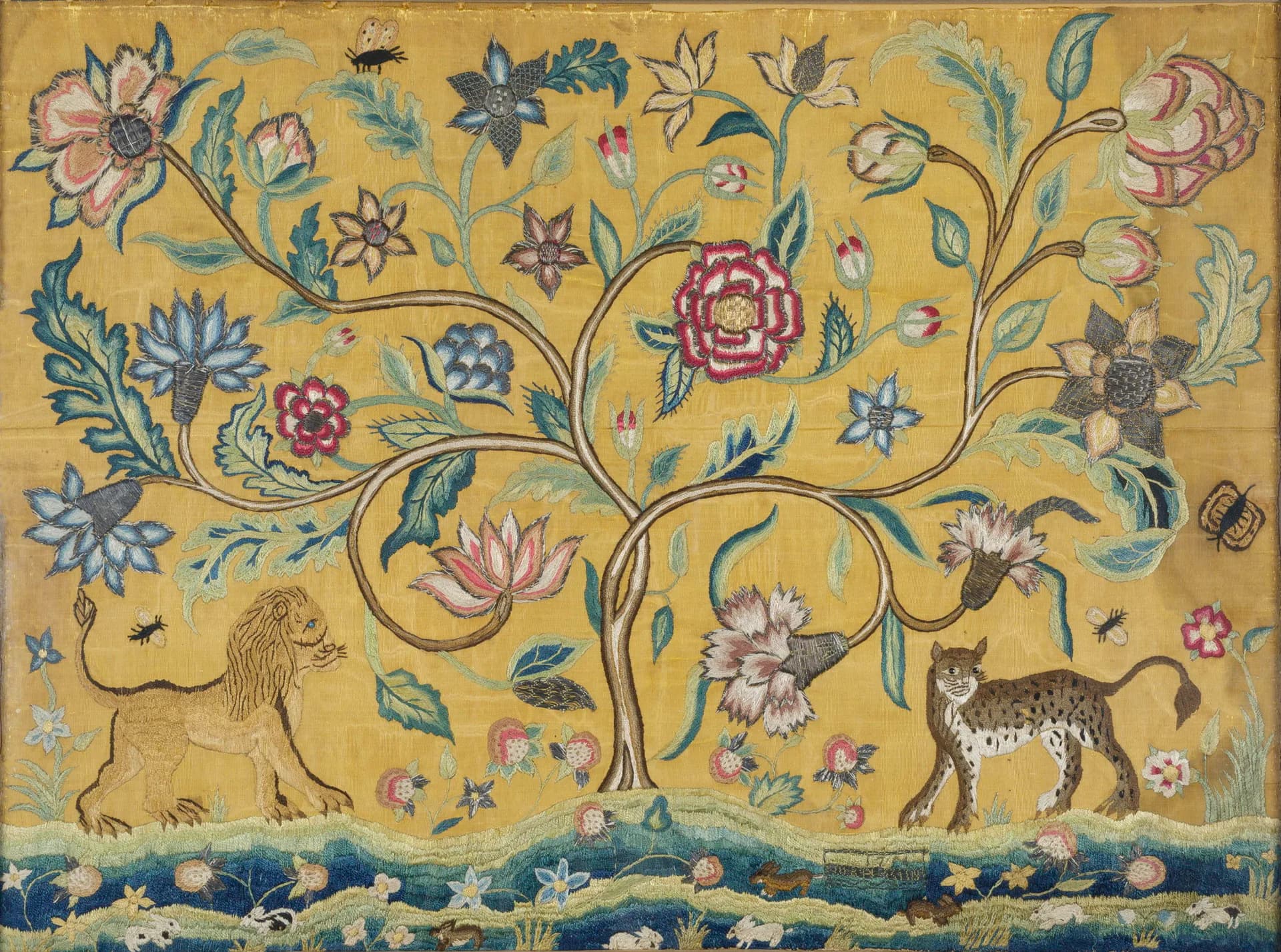Textile or embroidered design on golden background with flowering vines, animals including a dog and deer, and decorative border in blues and reds.