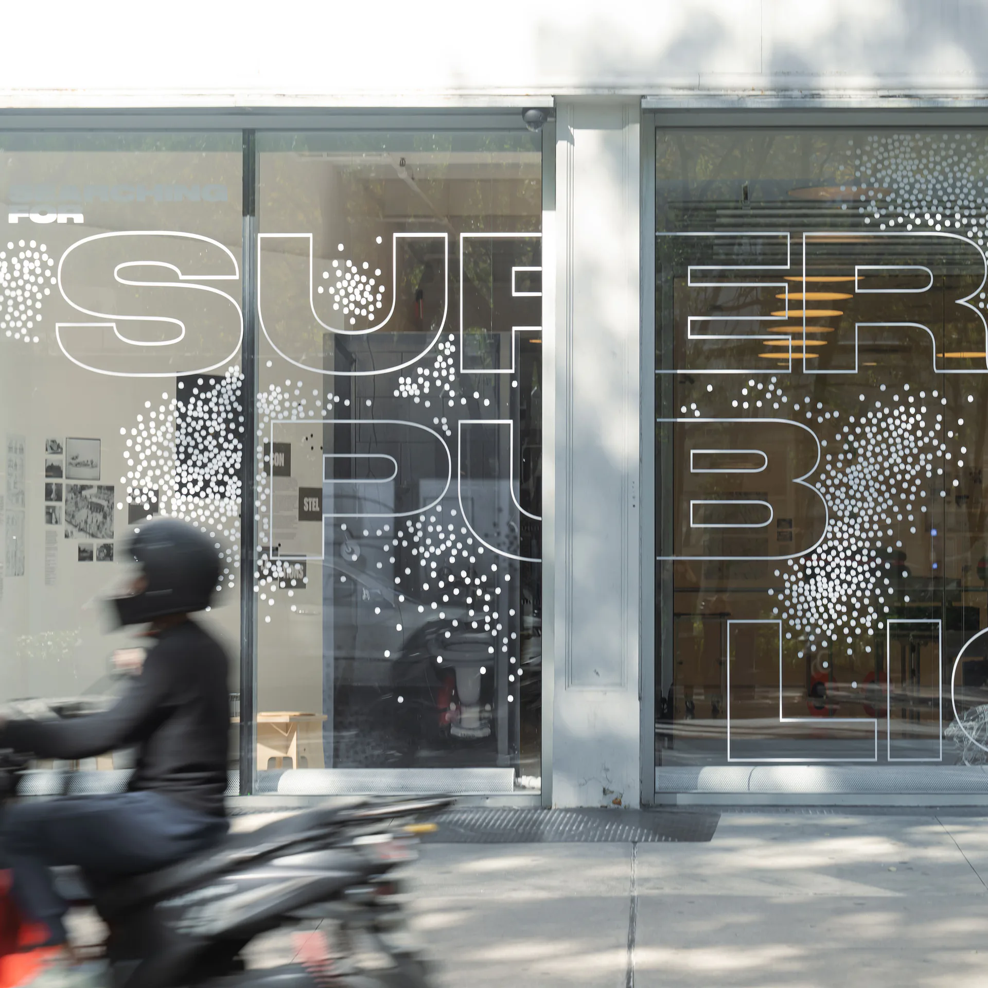 Modern storefront with glass doors and windows displaying large typography and dotted pattern design elements, pedestrian on motorcycle passing by.